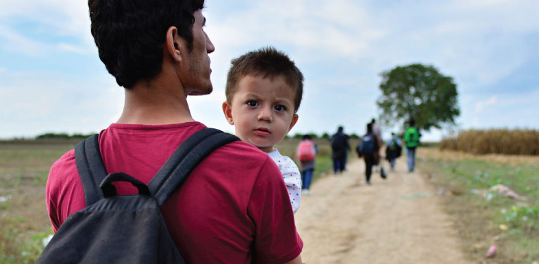 A group of people wearing backpacks walk down a path in a field. In the foreground, a parent holds a small child in his arms.