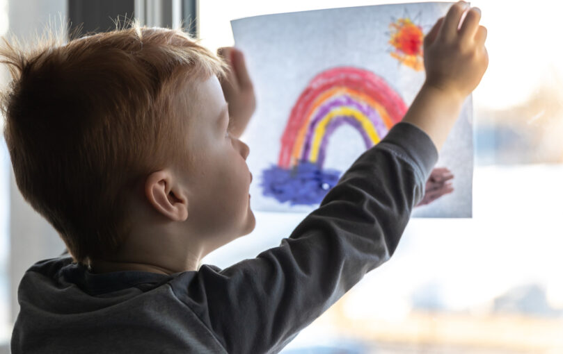 A young child placing his drawing of a rainbow and a sun in a window.
